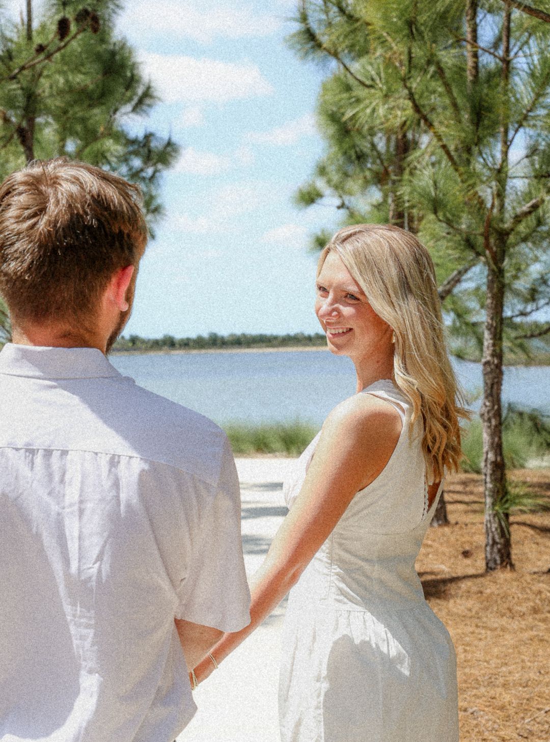 Blonde woman in white dress smiling at partner near pine trees by lake.