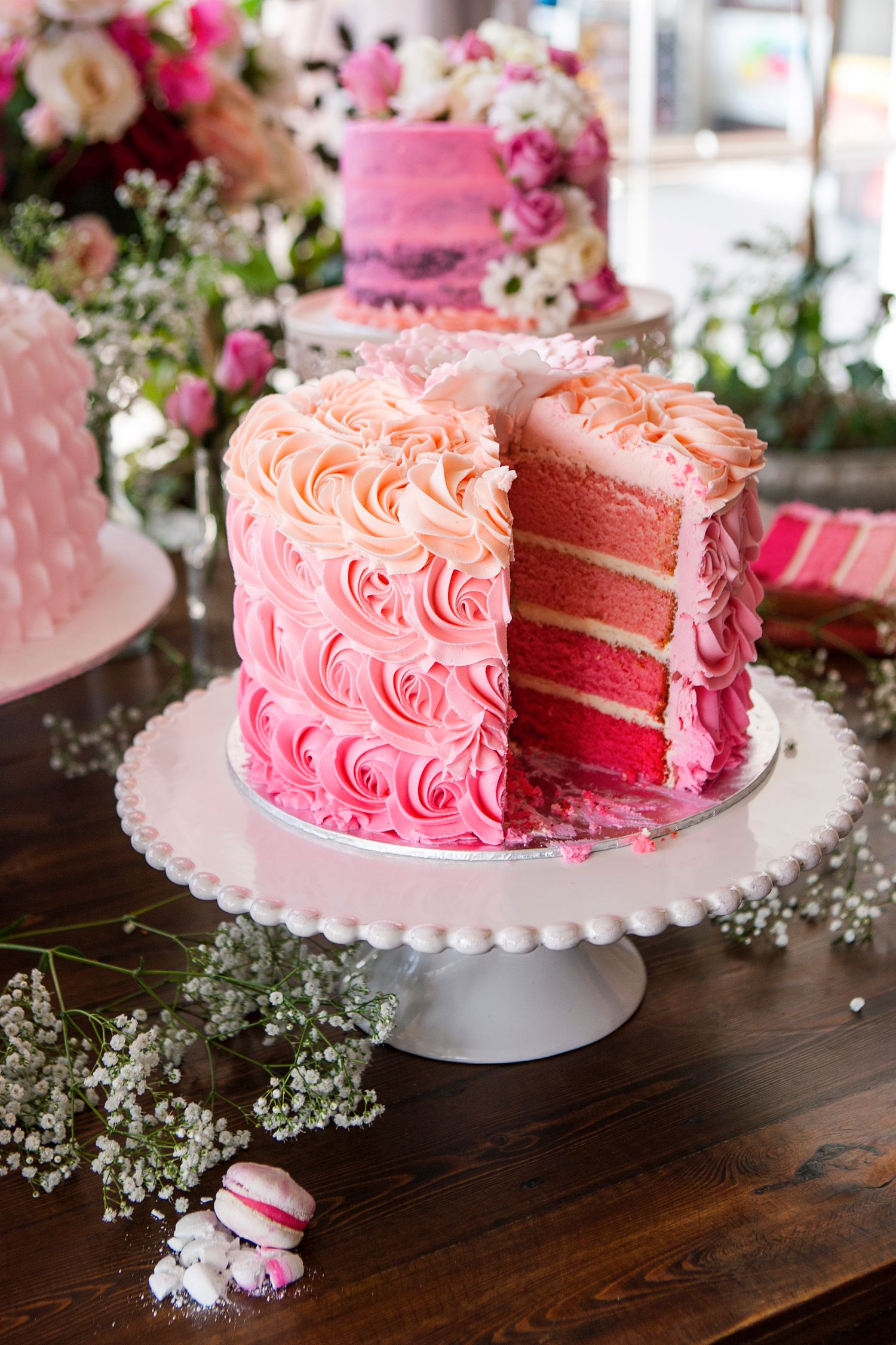 Sliced pink ombre rosette layer cake on white cake stand with macarons and flowers on wooden table.