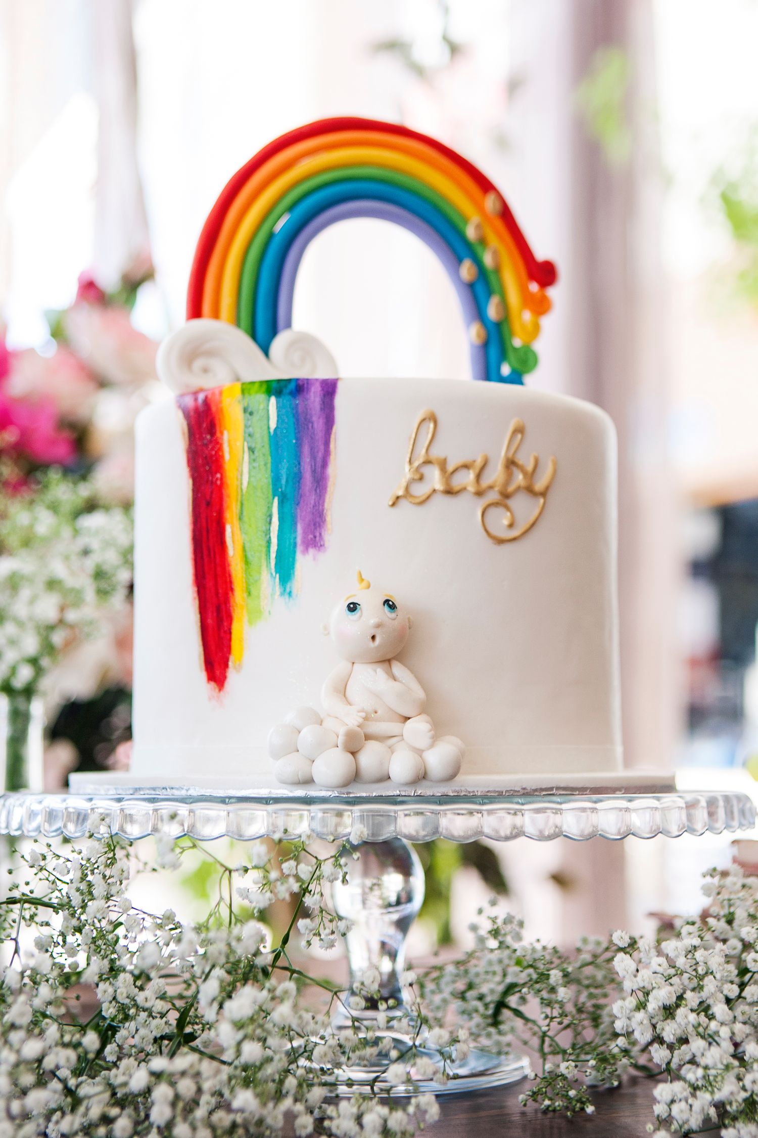 White baby shower cake with rainbow topper, unicorn figurine, and gold baby lettering on glass stand.