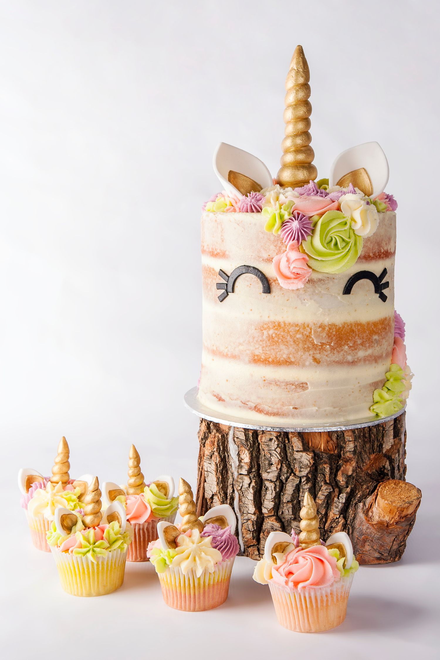 Unicorn cake on wood stump with gold horn, floral decorations, and matching unicorn cupcakes on white background.