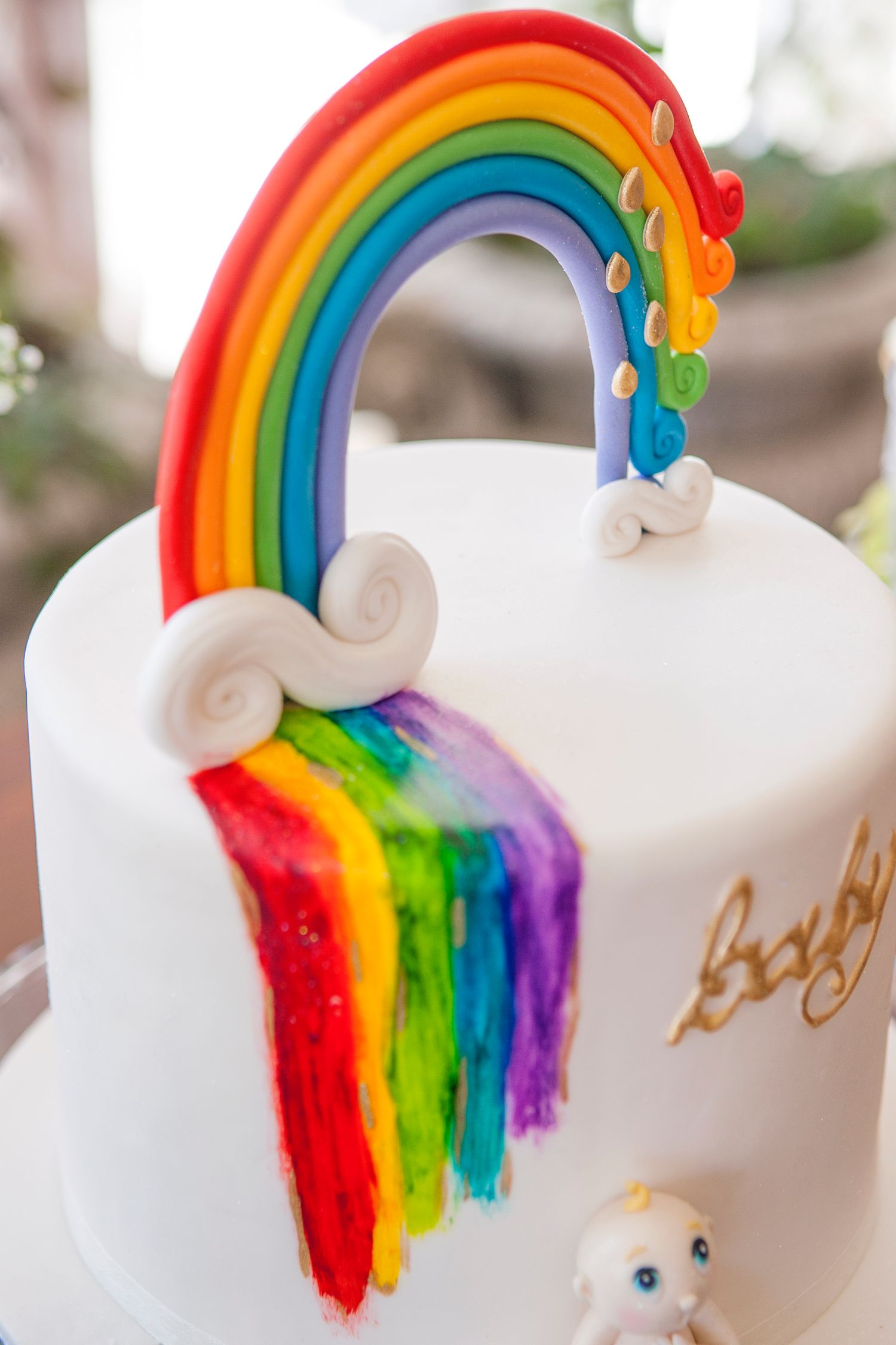 White baby shower cake with colorful rainbow fondant topper and painted rainbow drip design, featuring small baby figurine.