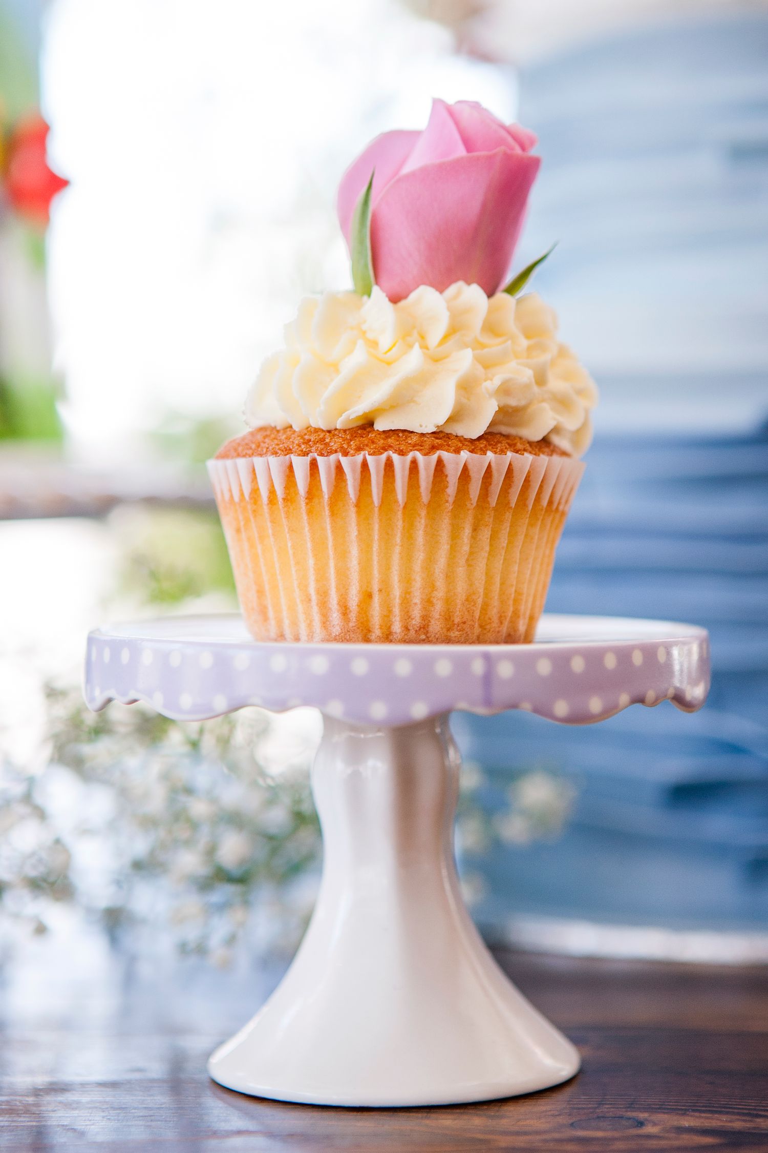 Vanilla cupcake with cream frosting and pink rose topper on polka dot lavender cake stand.
