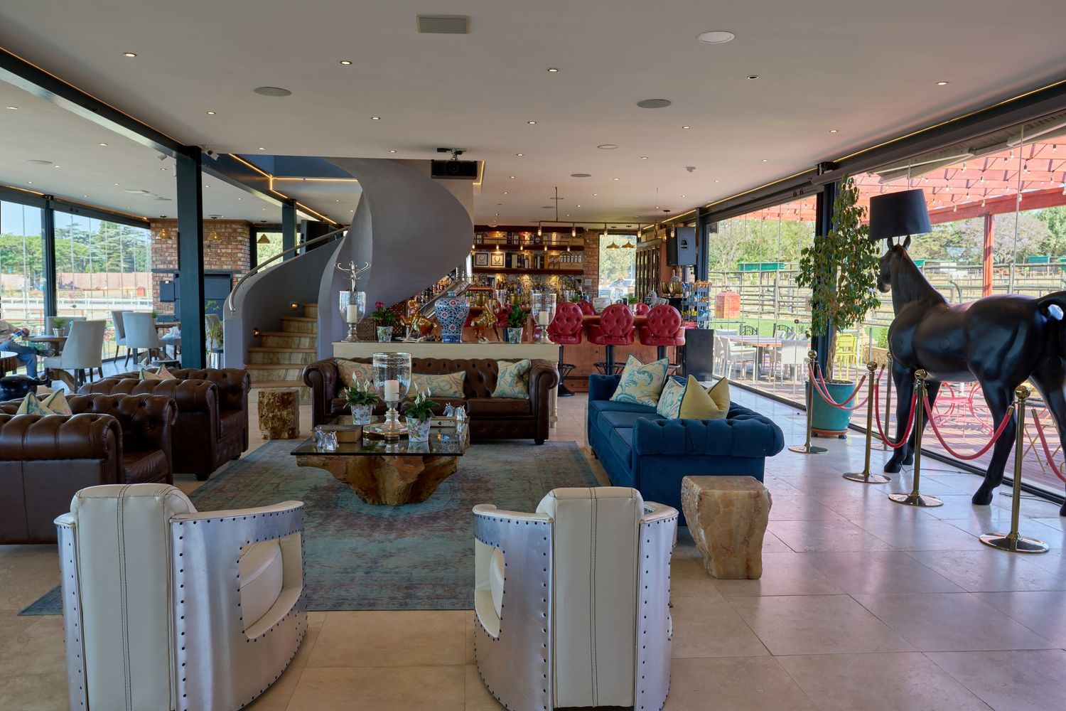 Elegant open-plan living space with eclectic furniture, bookshelf, spiral staircase, and decorative horse statue.