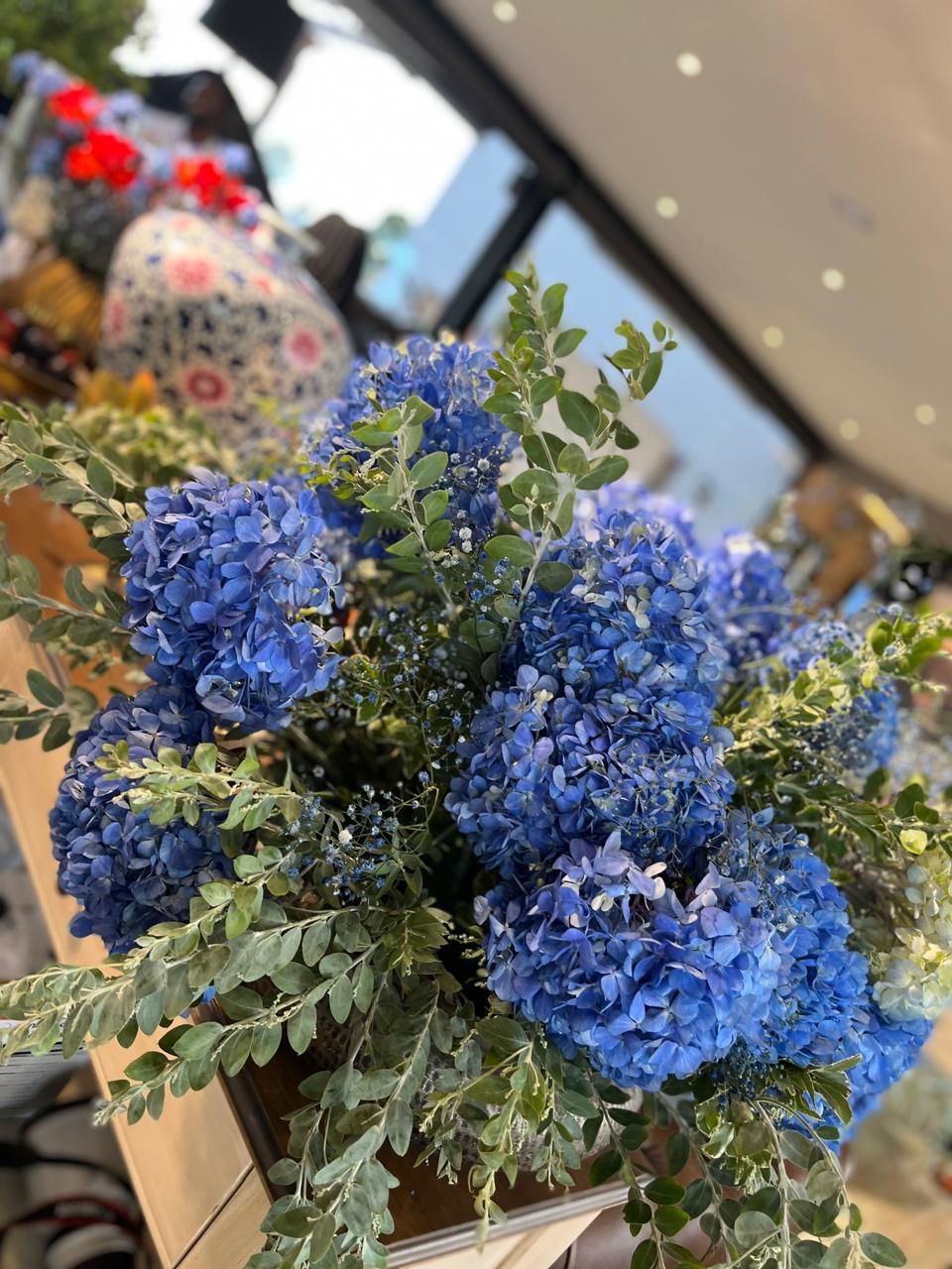 A close-up of a lush blue hydrangea and eucalyptus arrangement in a rustic container at an indoor floral event.