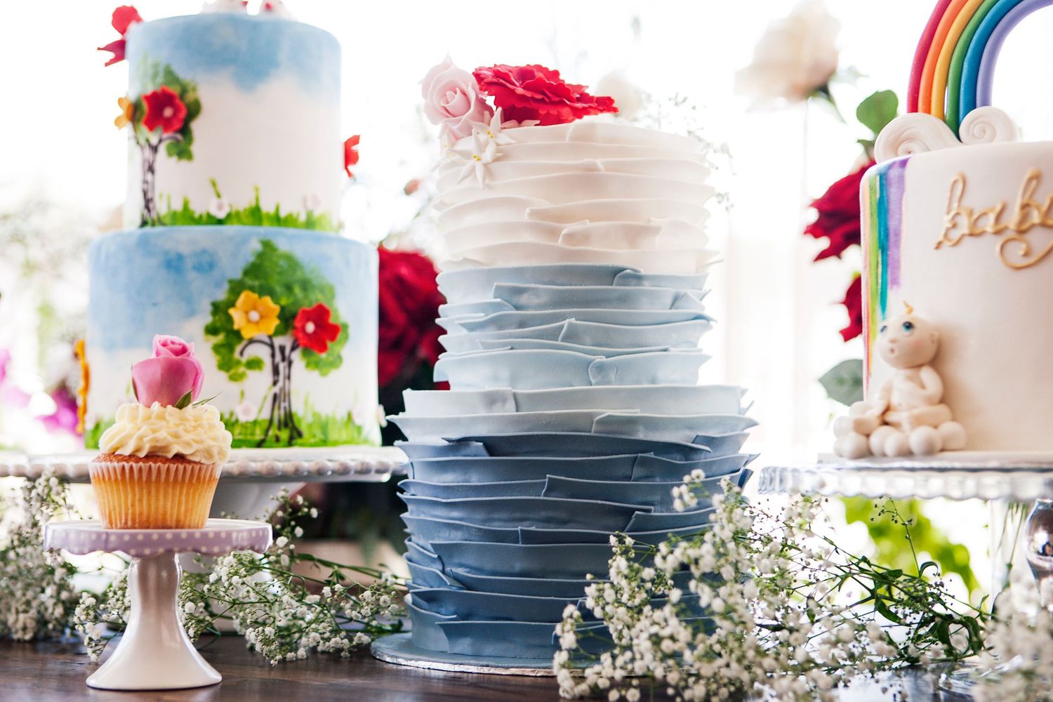 Elegant wedding cake display featuring ombre blue ruffled cake, floral painted cake, and rainbow cake with baby's breath.