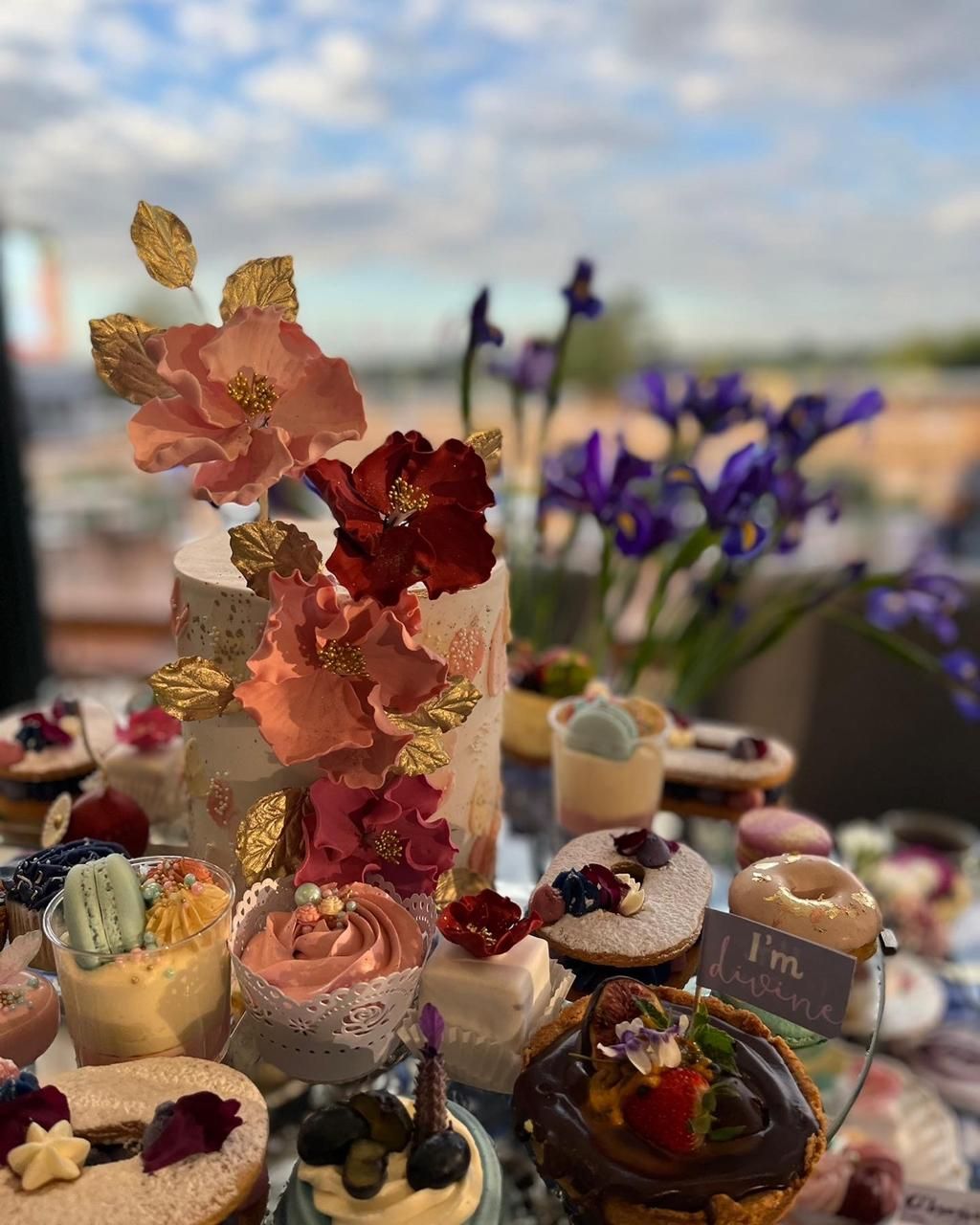 Elaborate dessert table featuring a floral-decorated cake surrounded by colorful cupcakes and pastries with a scenic sky backdrop.