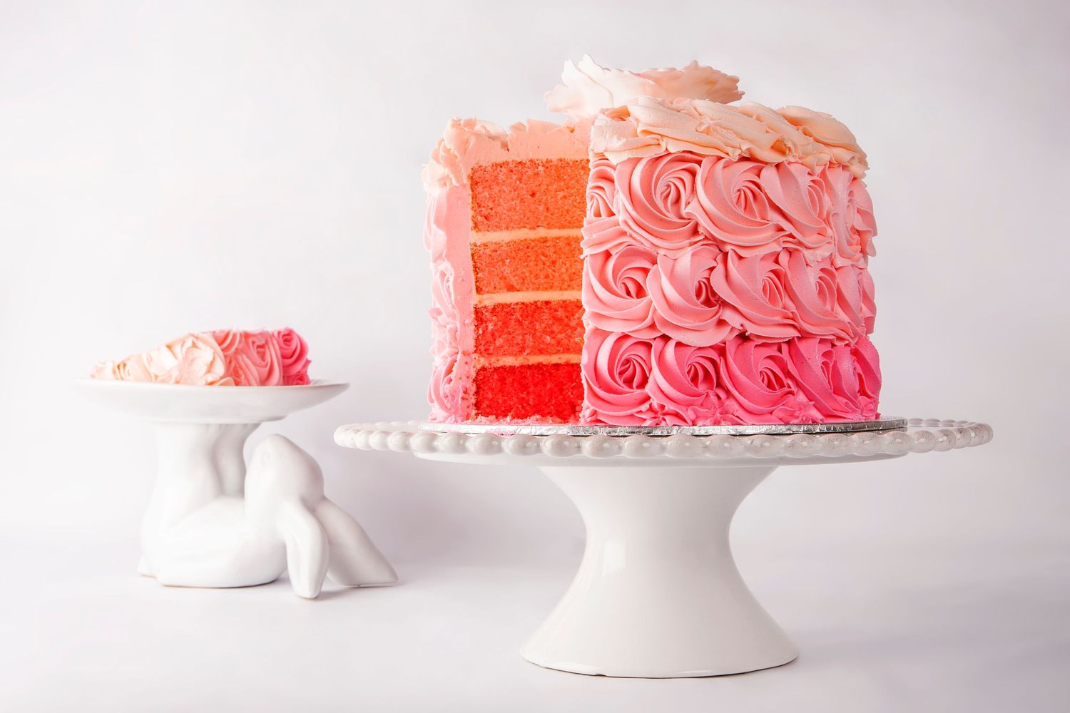 Pink ombre rosette layer cake on white cake stand with a slice served on small stand beside it.