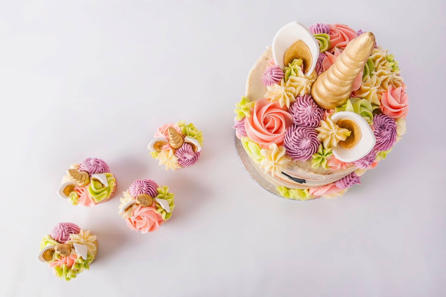 Top view of unicorn cake with colorful floral frosting, gold horn, and matching mini unicorn cupcakes.