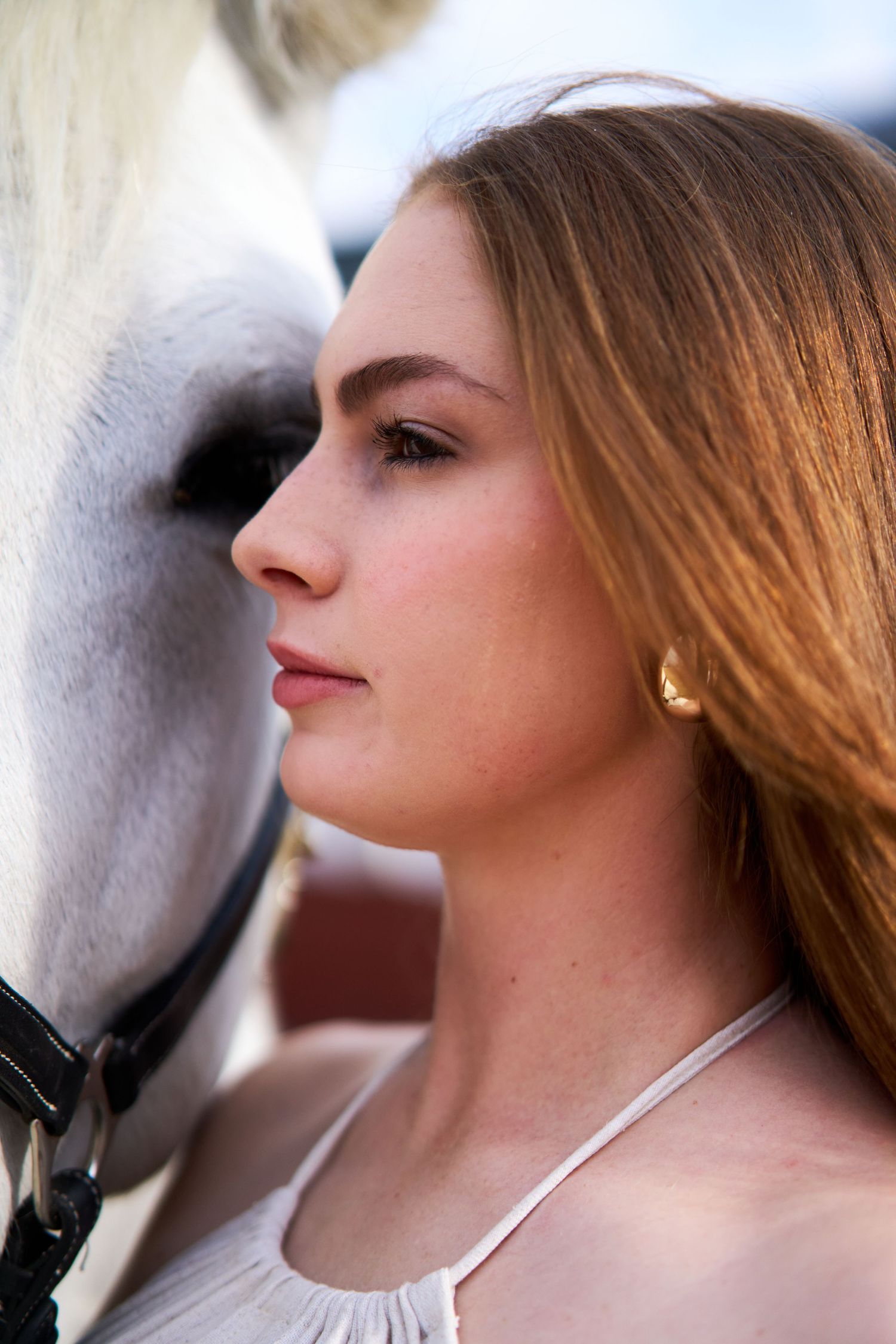 A close-up profile of a redheaded woman standing beside a white horse wearing a bridle.