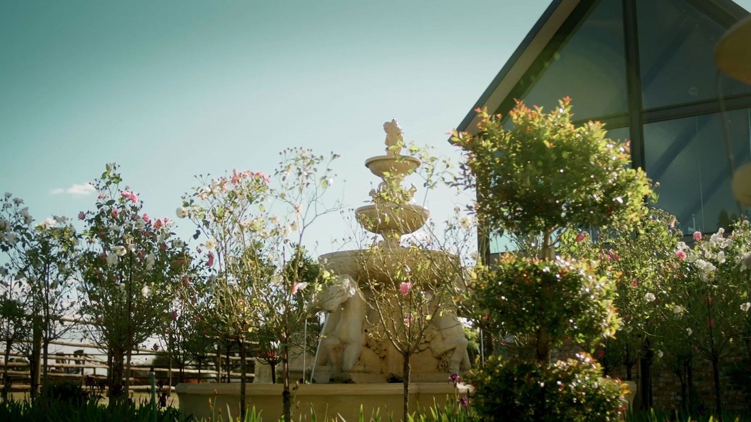 Ornate multi-tiered stone fountain surrounded by colorful flowering trees beside a modern glass building.