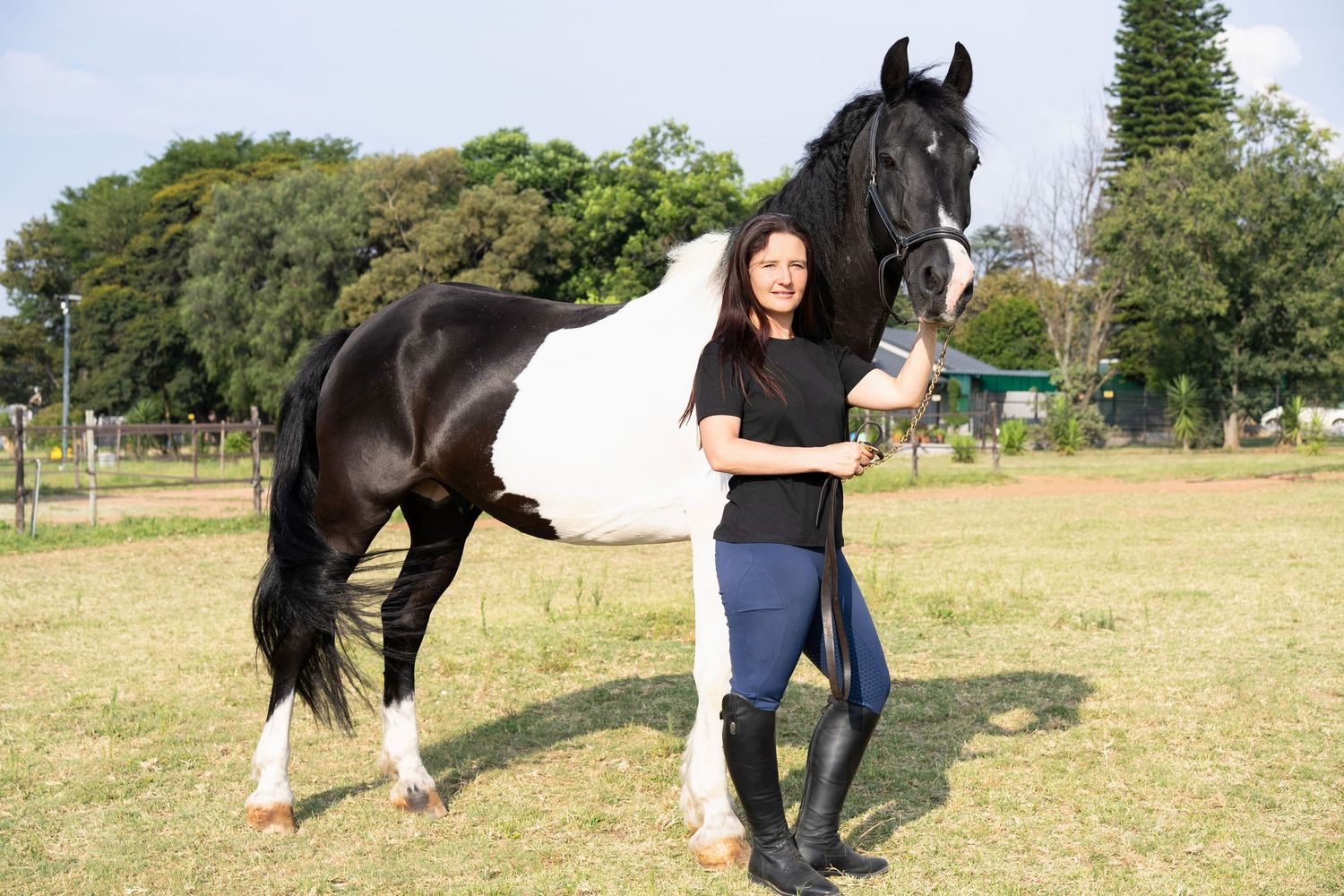 Woman in riding boots standing beside a black and white paint horse on green grass.