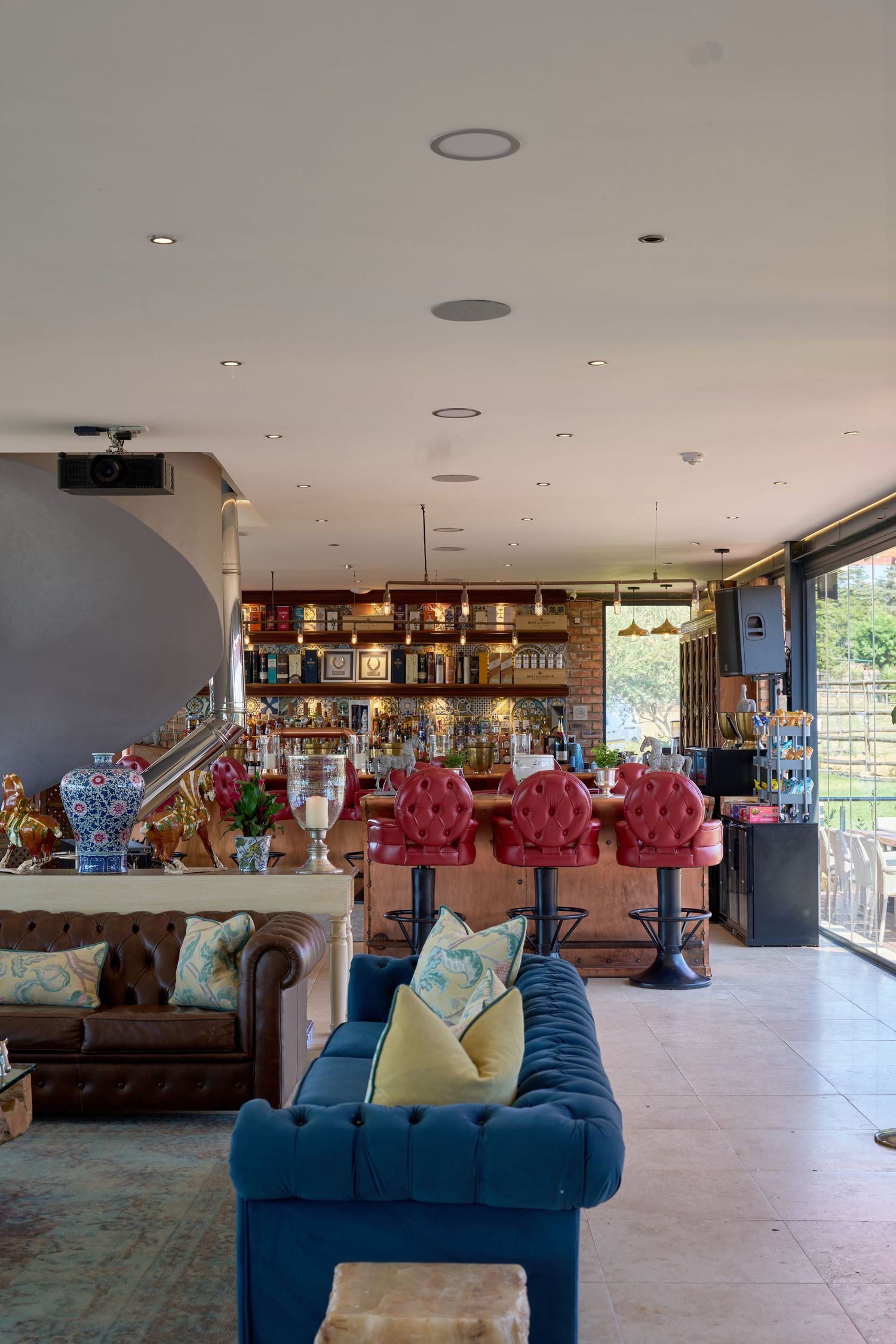 Stylish bar lounge featuring blue velvet sofa, red tufted bar stools, and well-stocked shelves with natural light.