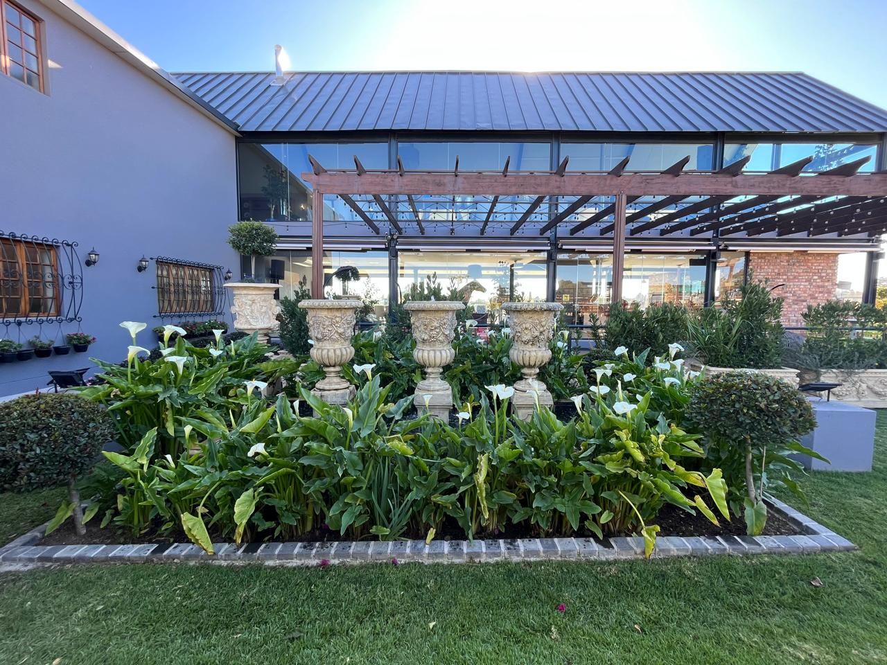 Outdoor venue garden bed featuring white calla lilies and ornate urns beside a modern glass structure.