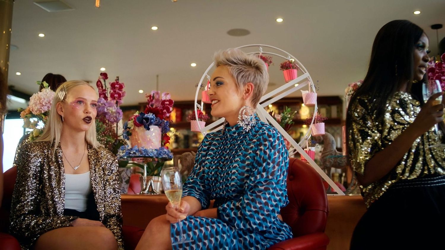 Three stylishly dressed women enjoying champagne in a vibrant, flower-decorated bar setting.