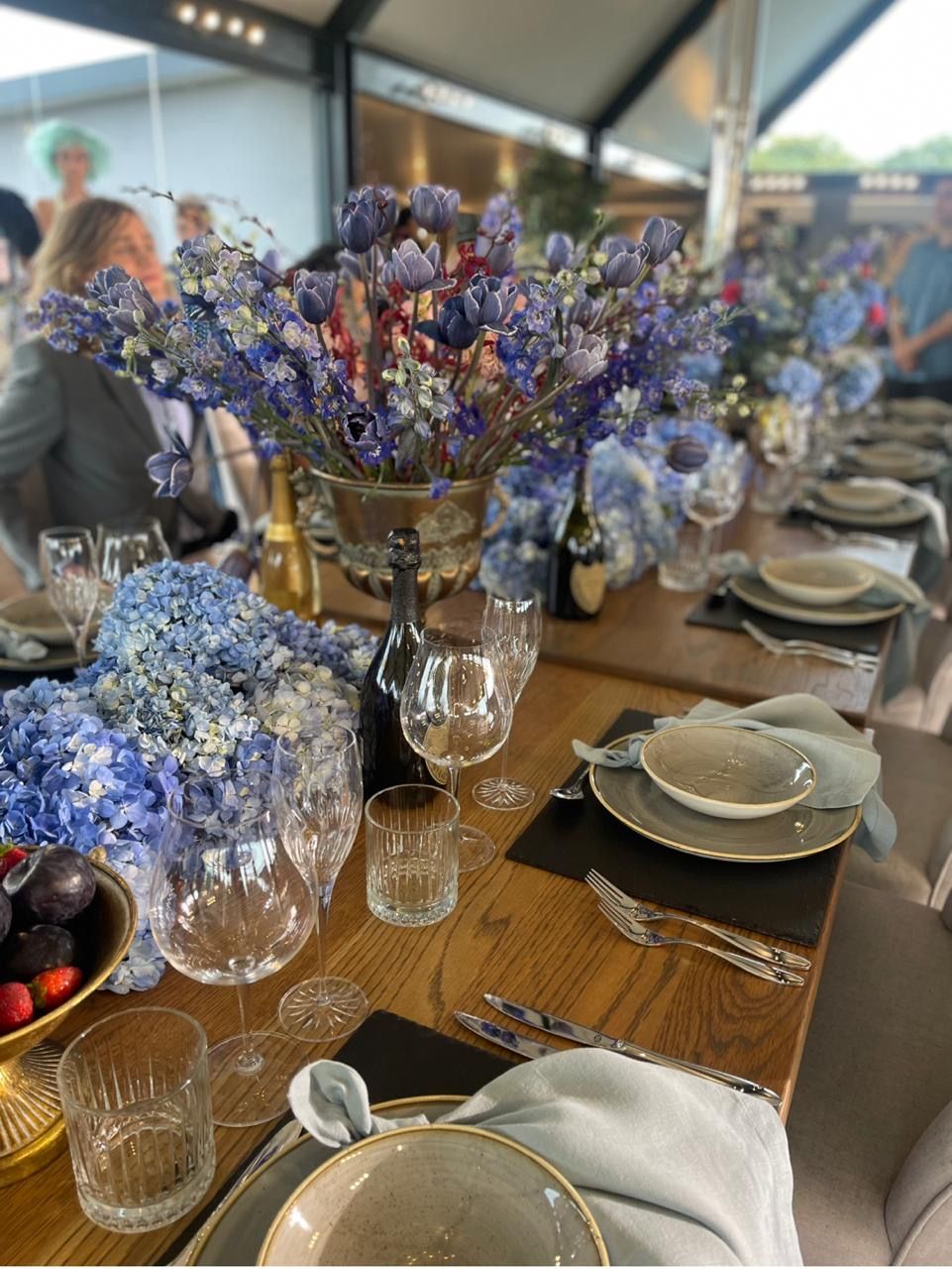 Long wooden dining table elegantly set with blue hydrangea centerpieces and crystal glassware for an event.