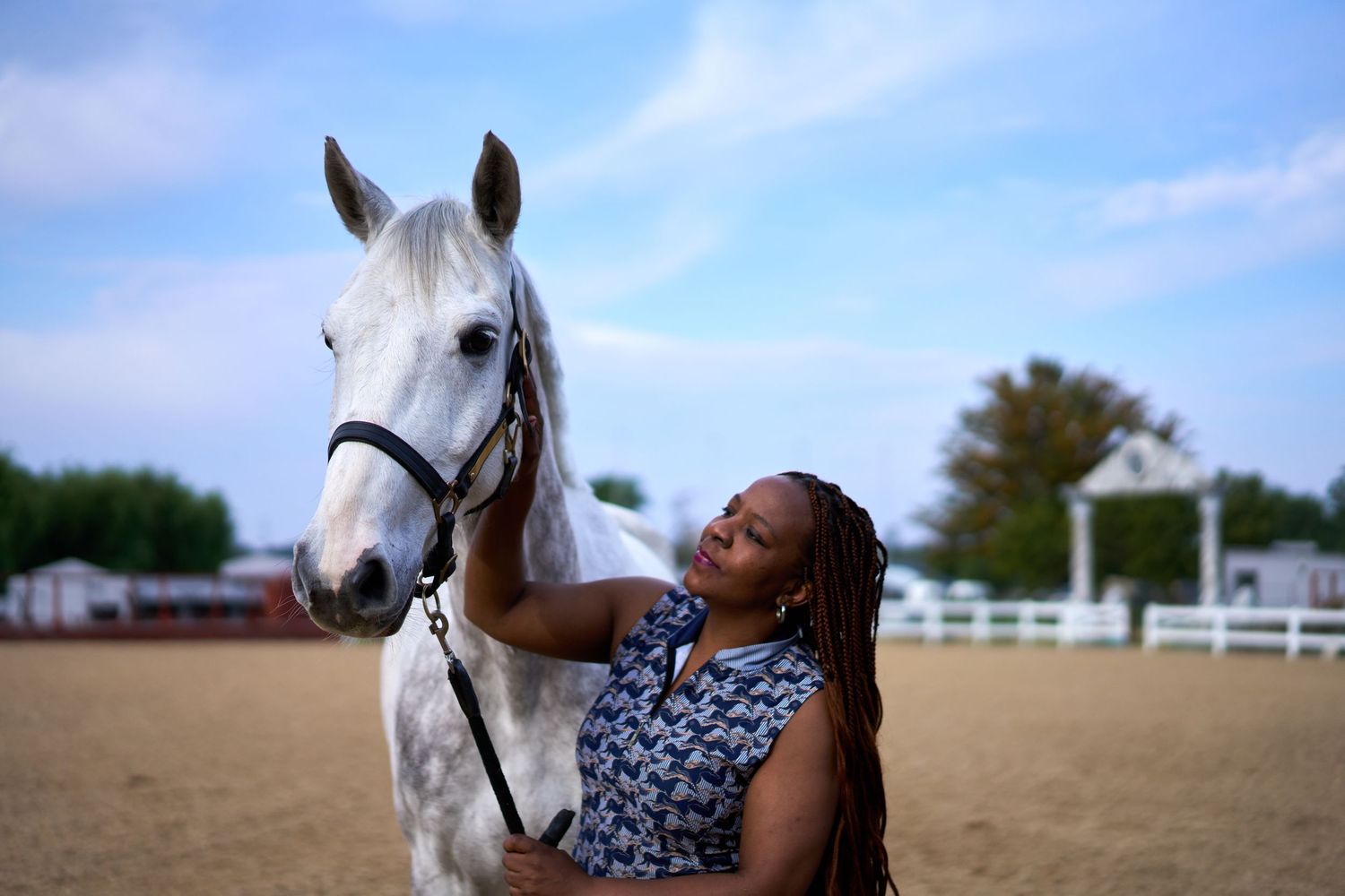 A woman with braids gently pets a dapple grey horse at an outdoor equestrian facility.