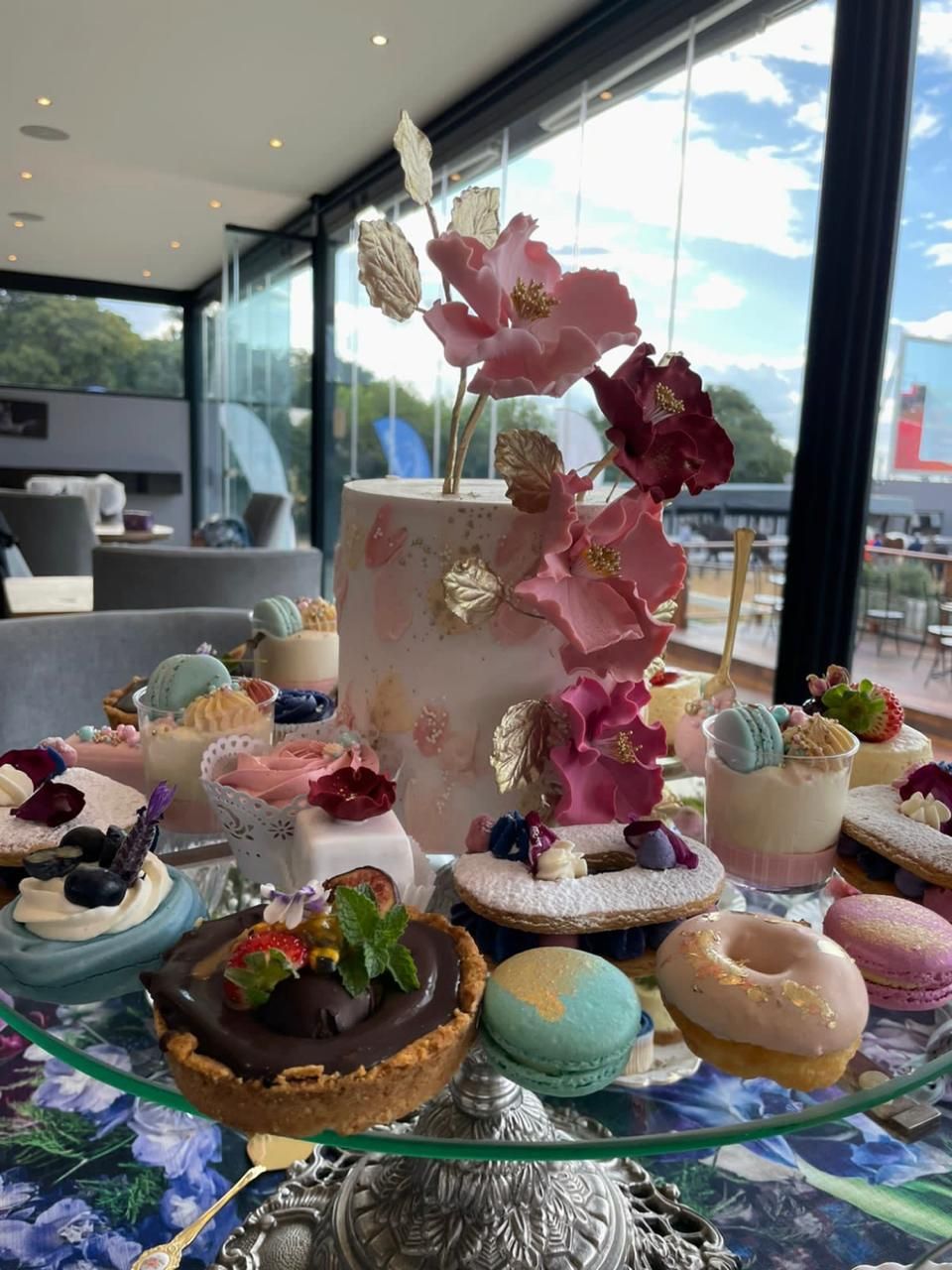 An elegant dessert table featuring a floral cake adorned with pink orchids, macarons, donuts, and tarts by a window.