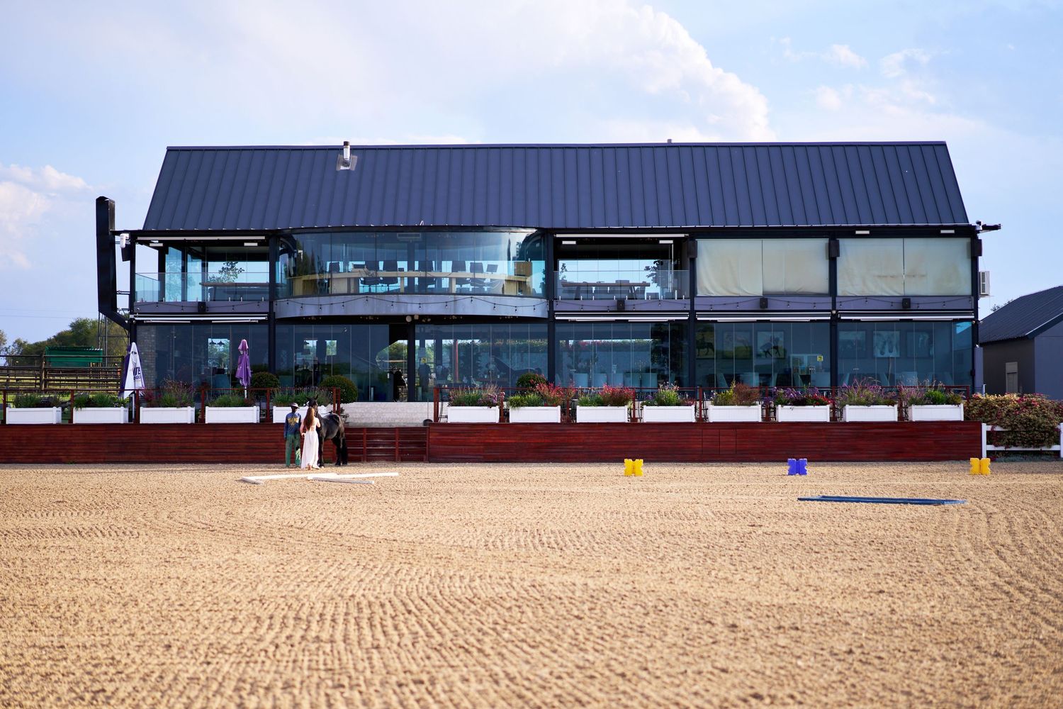 A modern equestrian arena building with large glass windows overlooking a sandy riding arena.