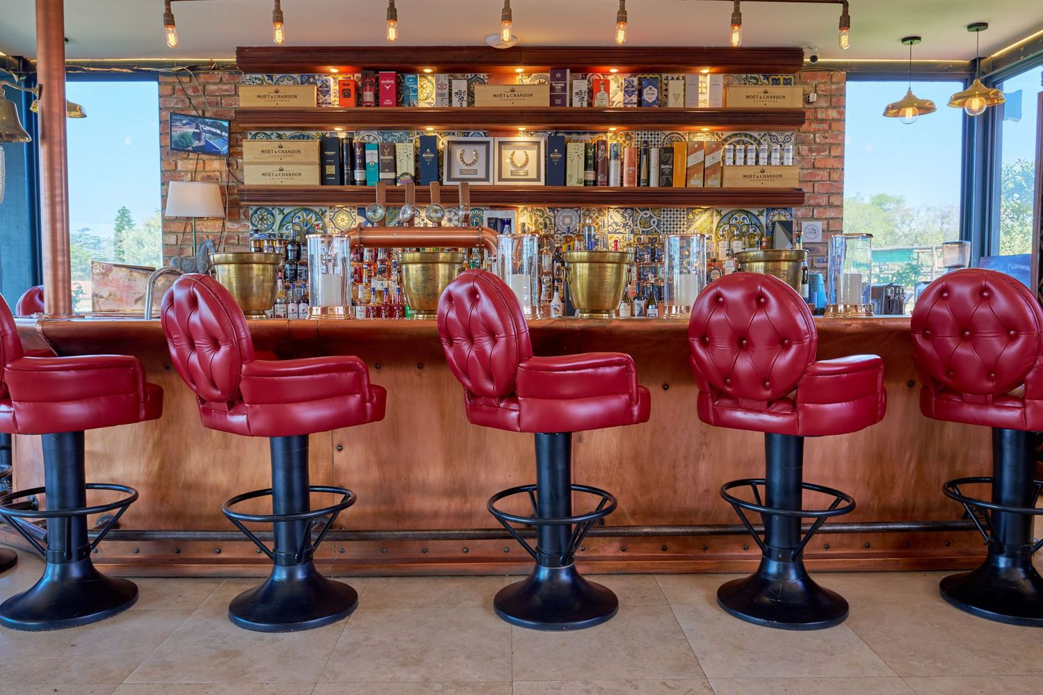 Stylish bar area with red tufted leather bar stools, a fully stocked wooden back bar, and city views.