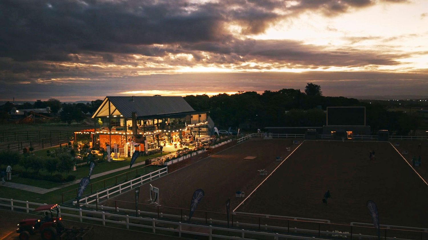 Aerial dusk view of a lit equestrian facility with riding arenas under a dramatic sunset sky.