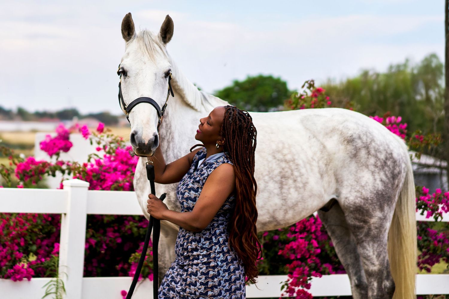A woman in a floral dress affectionately embraces a dapple grey horse near a white fence with pink flowers.