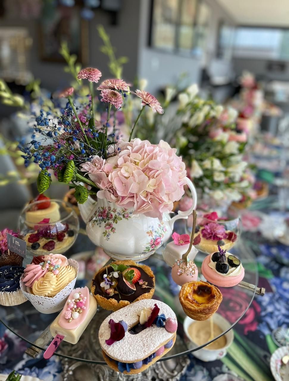 Vintage floral teapot with pink hydrangeas surrounded by elegant assorted pastries on a glass cake stand.