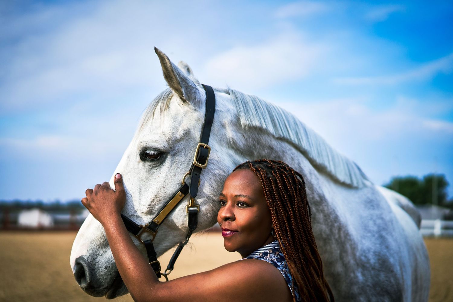Young woman with braids affectionately embracing a grey horse outdoors under a blue sky.