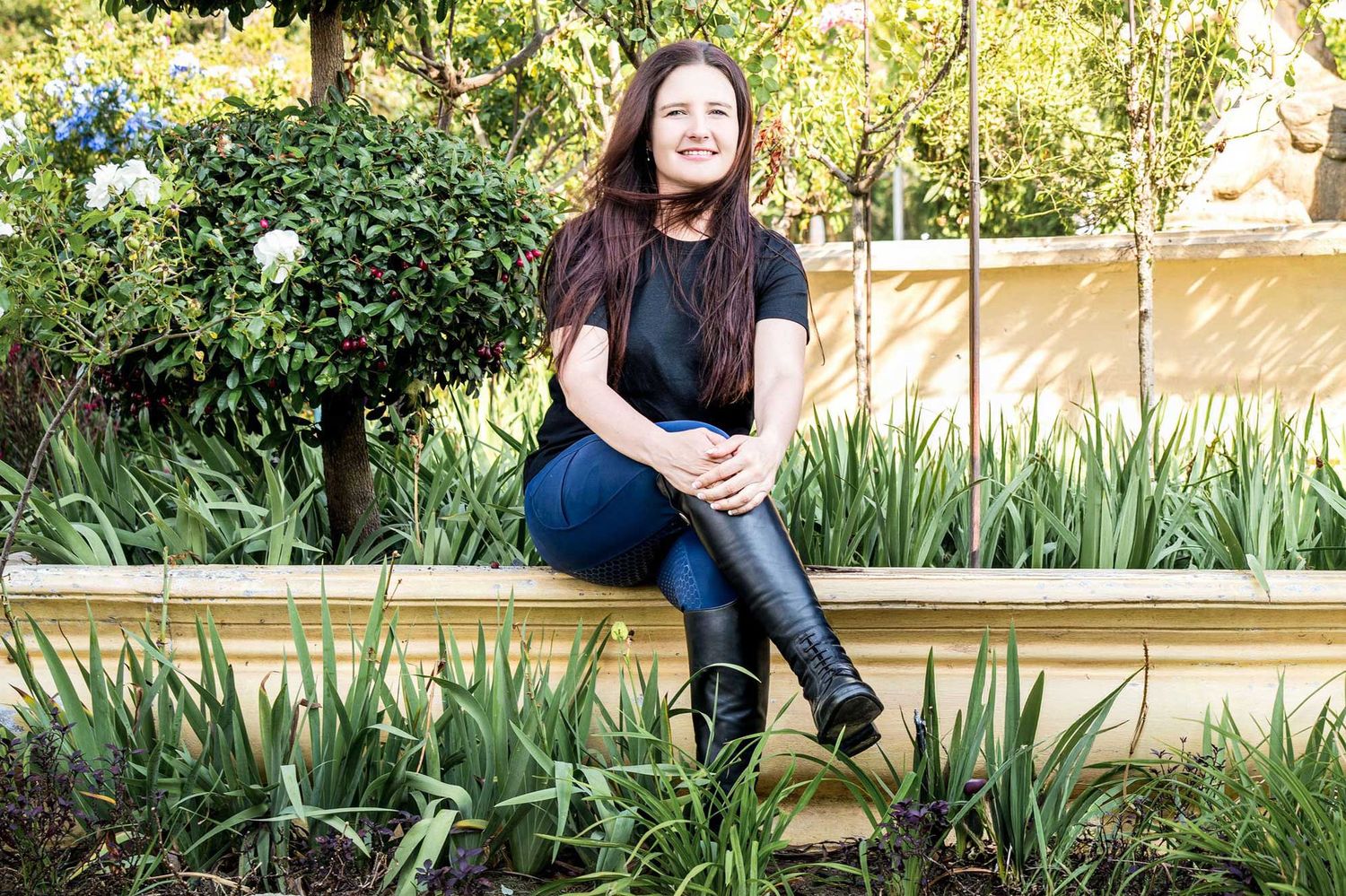 A smiling woman with long dark hair sits on a garden planter surrounded by lush green plants and flowers.