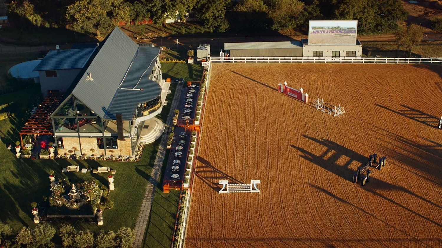 Aerial view of an equestrian facility with riding arena and stable buildings at sunset.