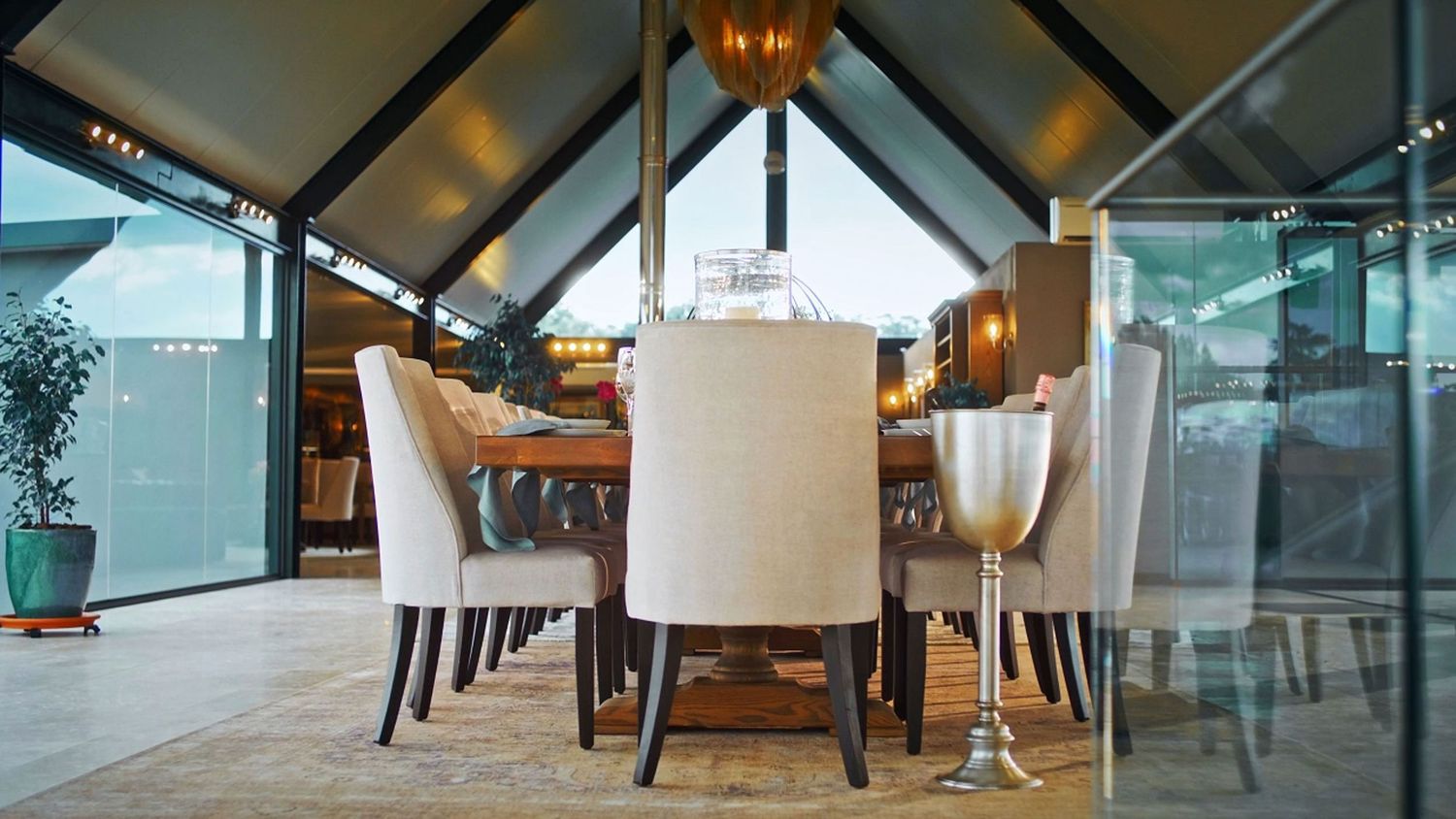 Sophisticated dining area with upholstered chairs, wooden table, and dramatic angled glass ceiling architecture.