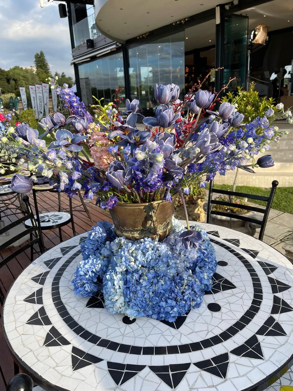 A vibrant arrangement of blue and purple flowers including tulips and hydrangeas in a gold vase on a mosaic table.
