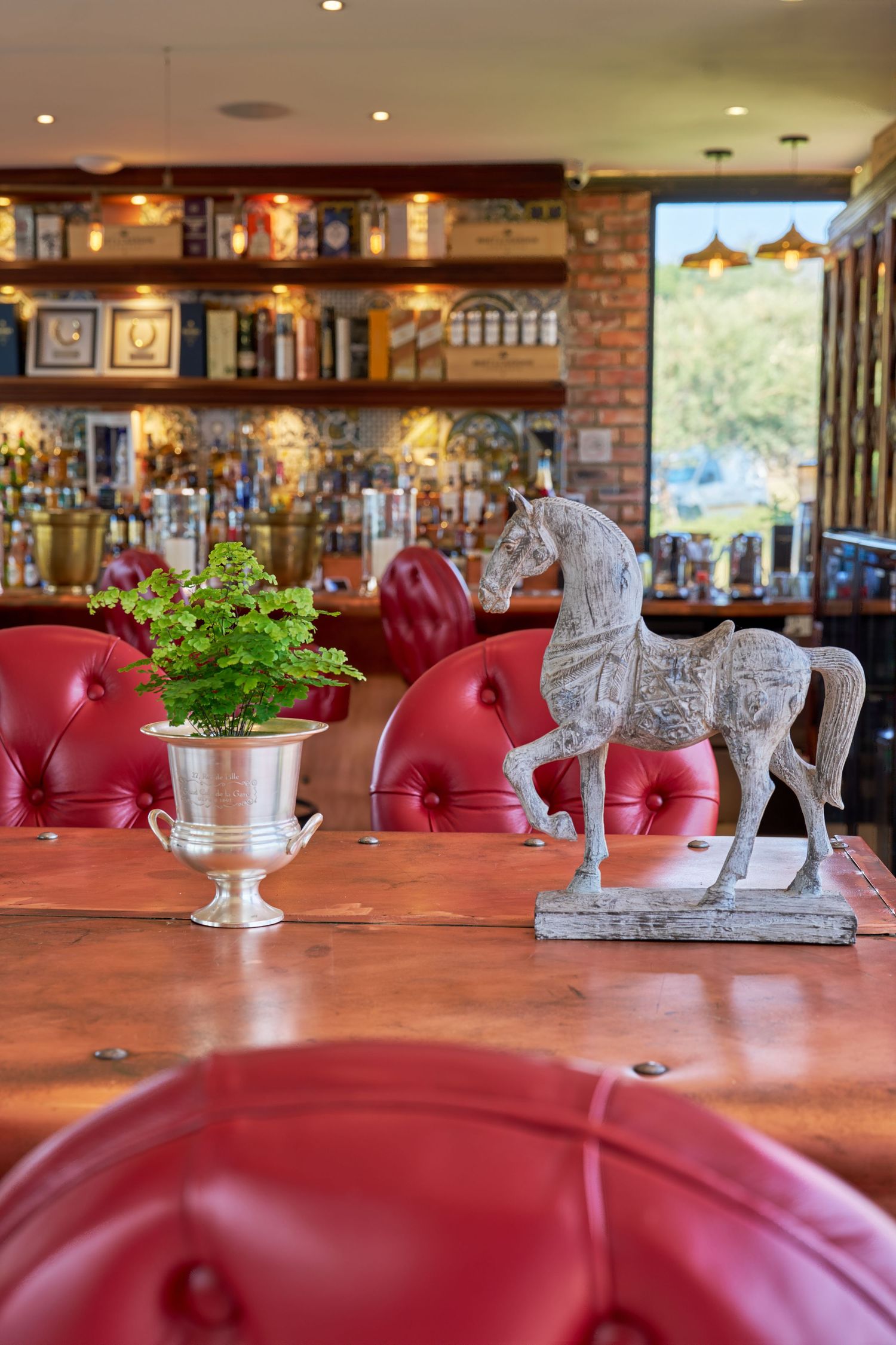 Rustic bar interior with red leather chairs, wooden table, horse statue decor, and backlit liquor shelves.