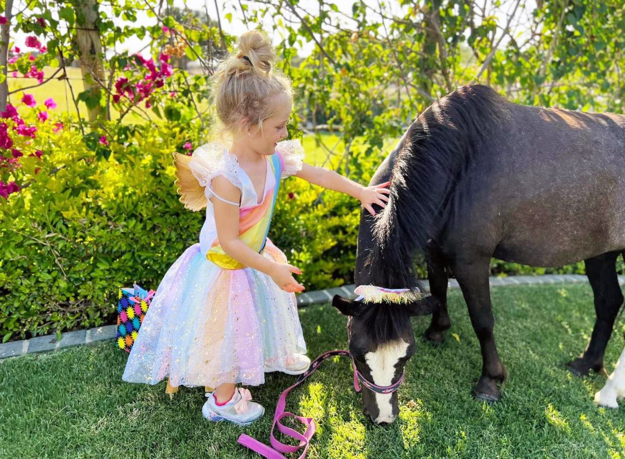 Young girl in rainbow fairy dress petting a small black pony in a lush garden.