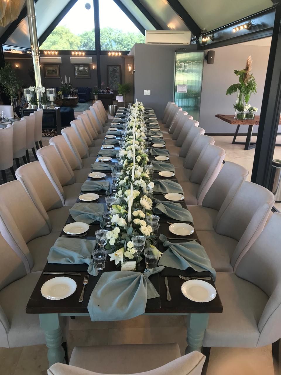 Formal long dining table with sage green napkins, white roses, and neutral upholstered chairs in a venue.