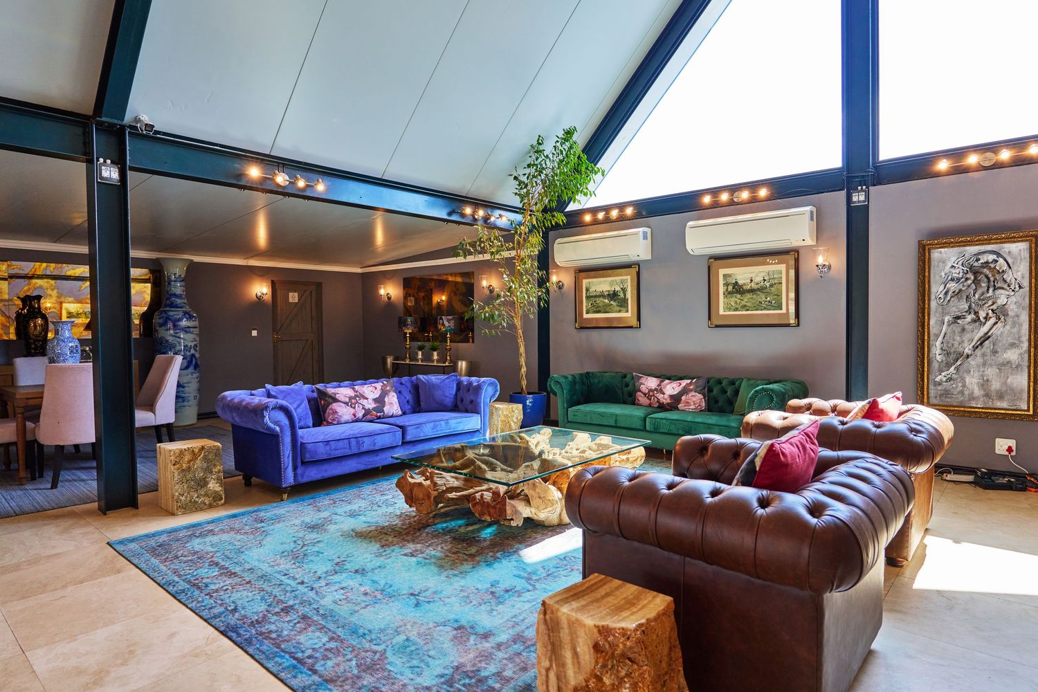 Luxurious living room with vibrant blue and green velvet sofas, leather couch, colorful rug, and high vaulted glass ceiling.