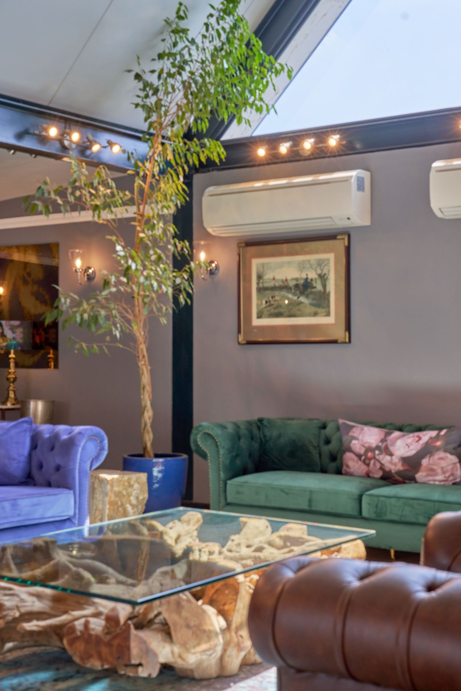 Elegant lounge with green velvet Chesterfield sofa, blue armchair, glass coffee table, indoor tree, and wall-mounted air conditioner.
