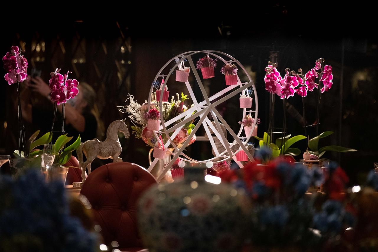 A decorative miniature Ferris wheel centerpiece surrounded by pink orchids and colorful floral arrangements at night.