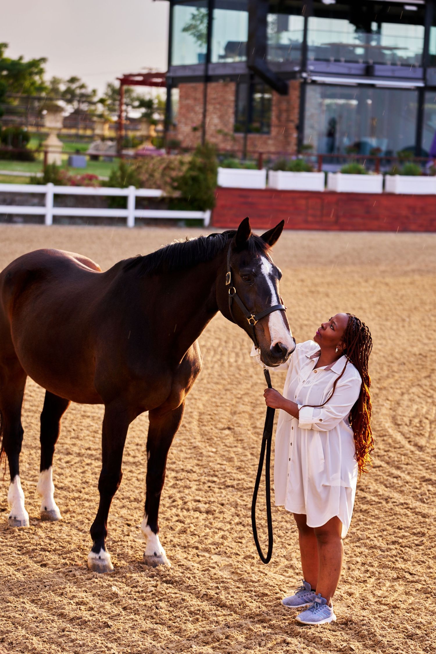 A woman in a white dress smiles while standing beside a bay horse in a sandy equestrian arena.