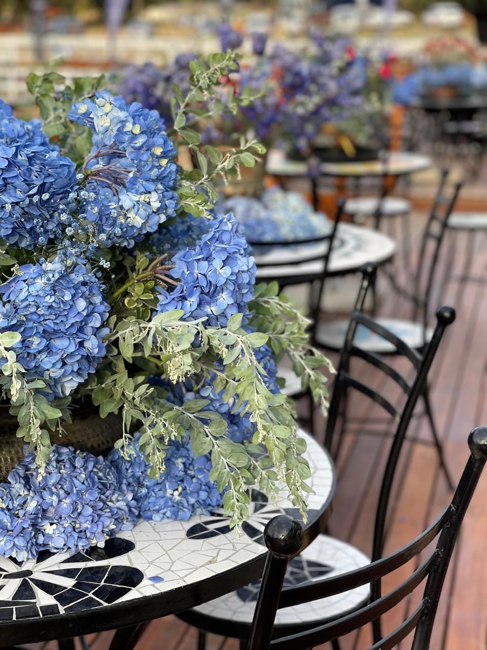 Blue hydrangea centerpieces on black and white mosaic bistro tables on an outdoor terrace with wrought iron chairs.