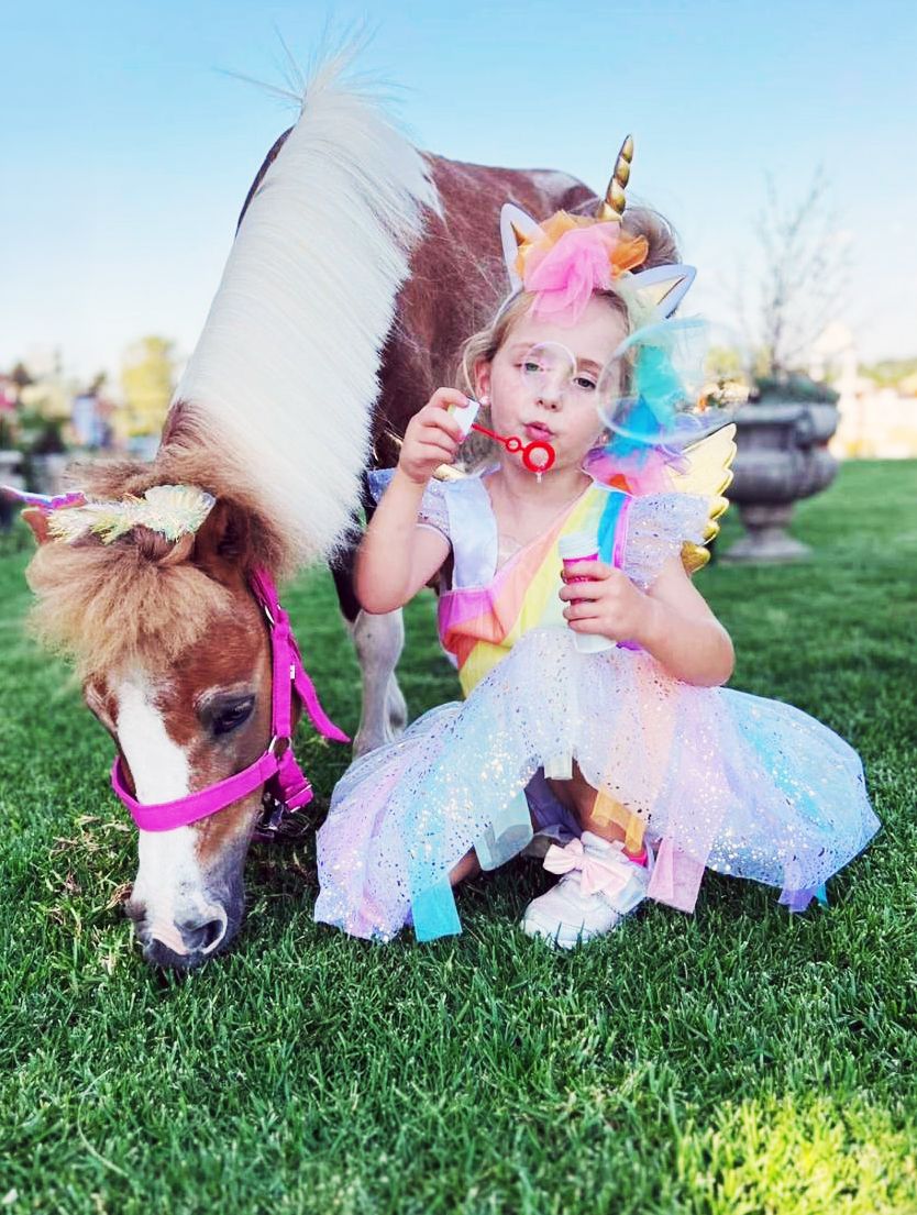 Girl in unicorn costume blowing bubbles beside a decorated mini pony on green grass.