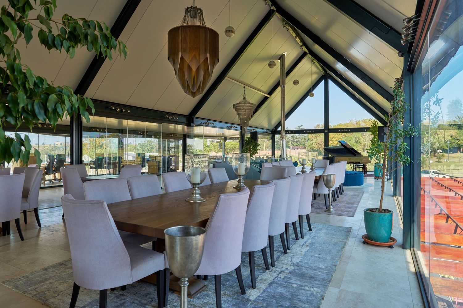 Spacious glass-enclosed dining room with a long wooden table, white upholstered chairs, and panoramic garden views.