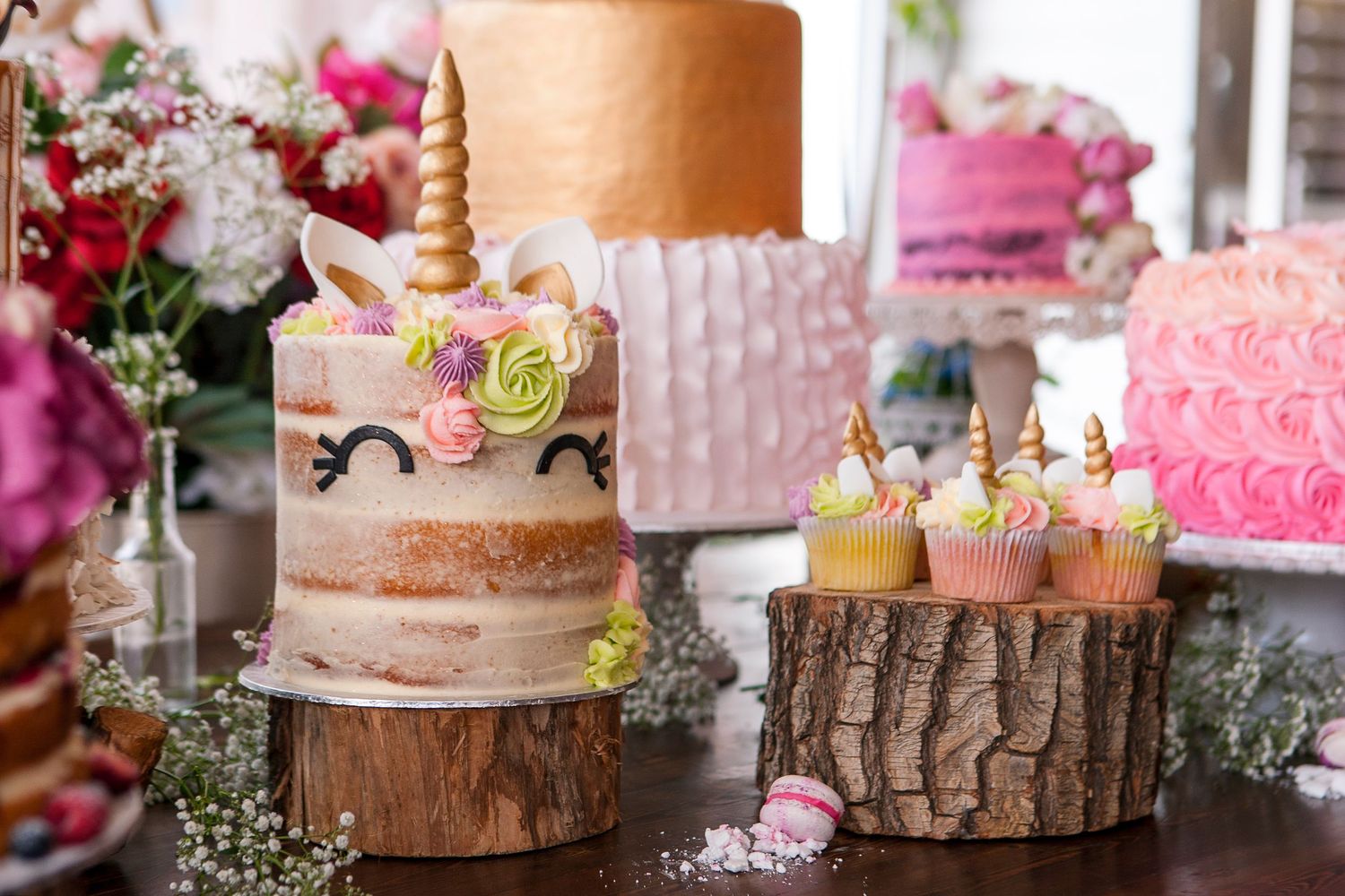 Unicorn cake with gold horn and floral decorations displayed on wooden stumps with cupcakes at a dessert table.