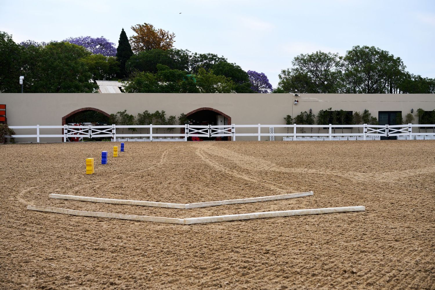 An empty sand equestrian arena with white fencing and colorful jump poles on a sunny day.