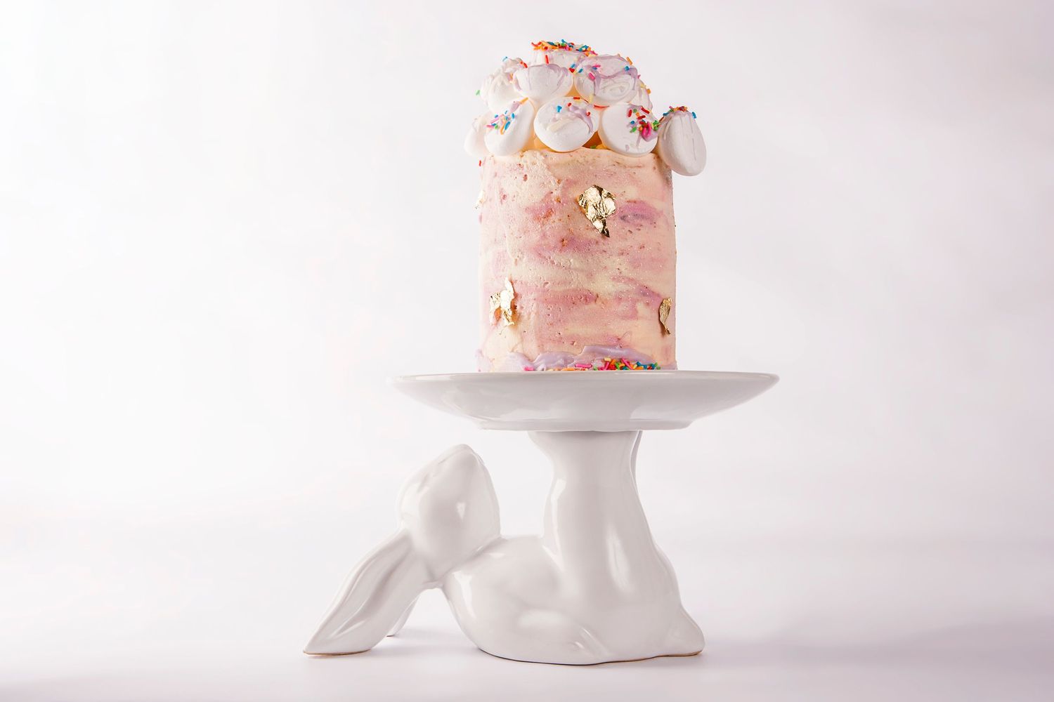 Pink marbled celebration cake with gold accents displayed on white sculptural angel cake stand.