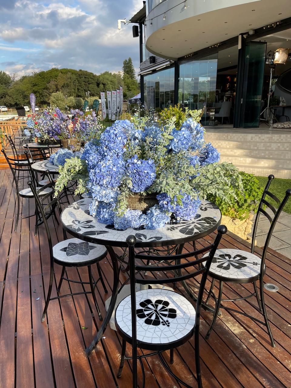 Blue hydrangea floral centerpiece on mosaic table on a wooden deck outdoor dining area.