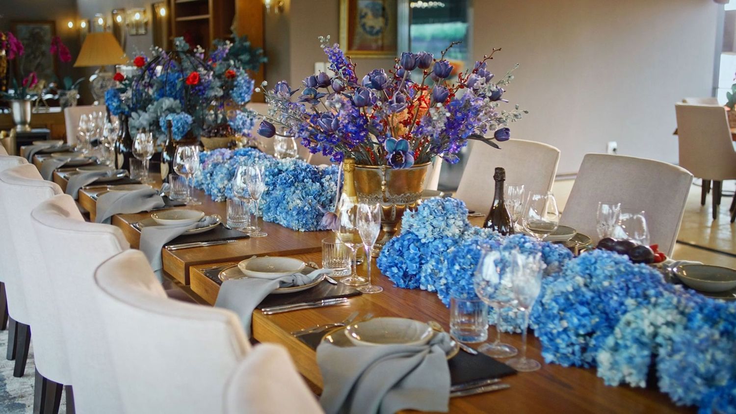 Elegant long dining table adorned with blue hydrangea centerpieces and purple floral arrangements in gold vases.