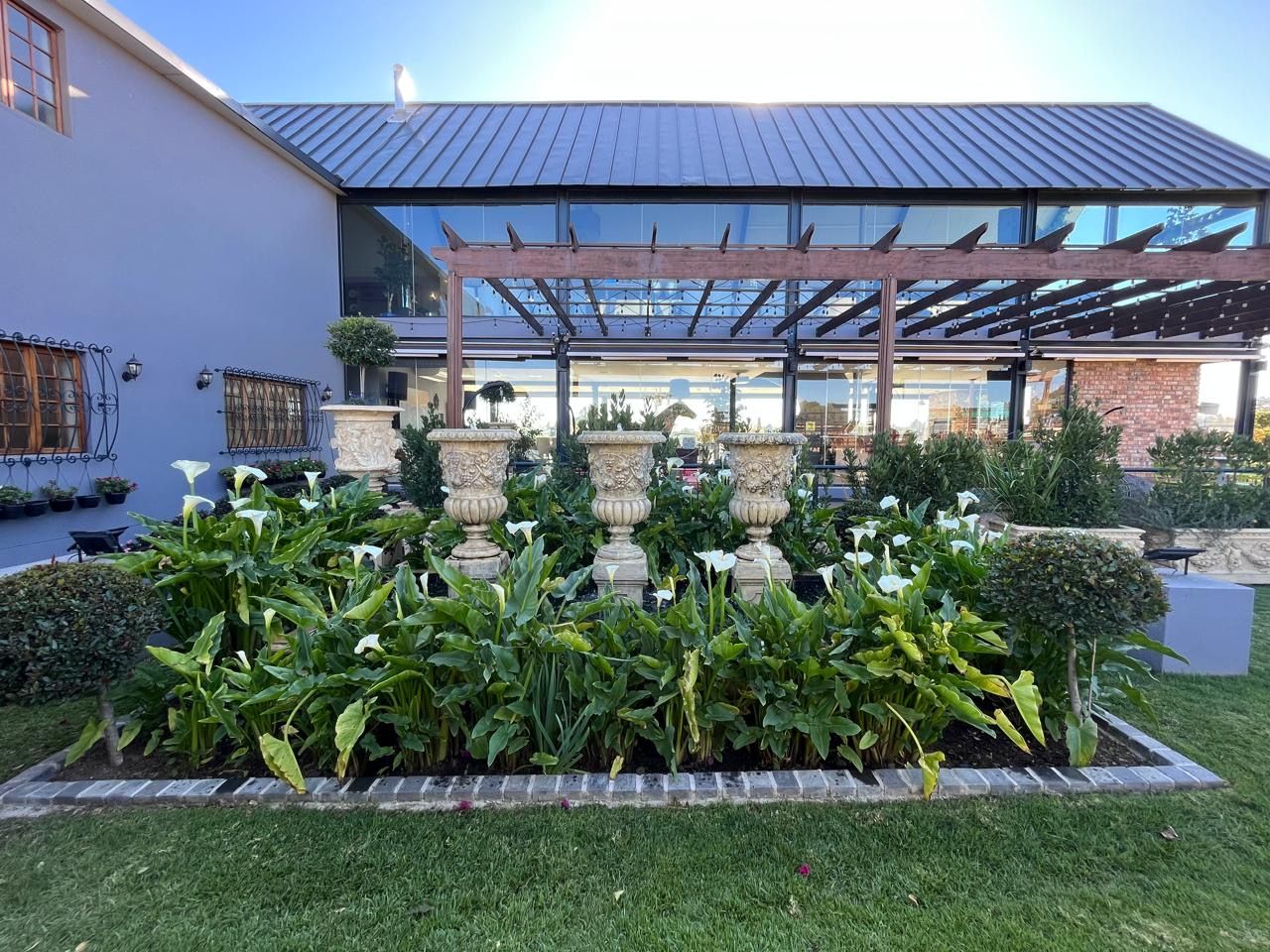 Outdoor venue garden bed featuring white calla lilies and ornate urns beside a modern glass structure.