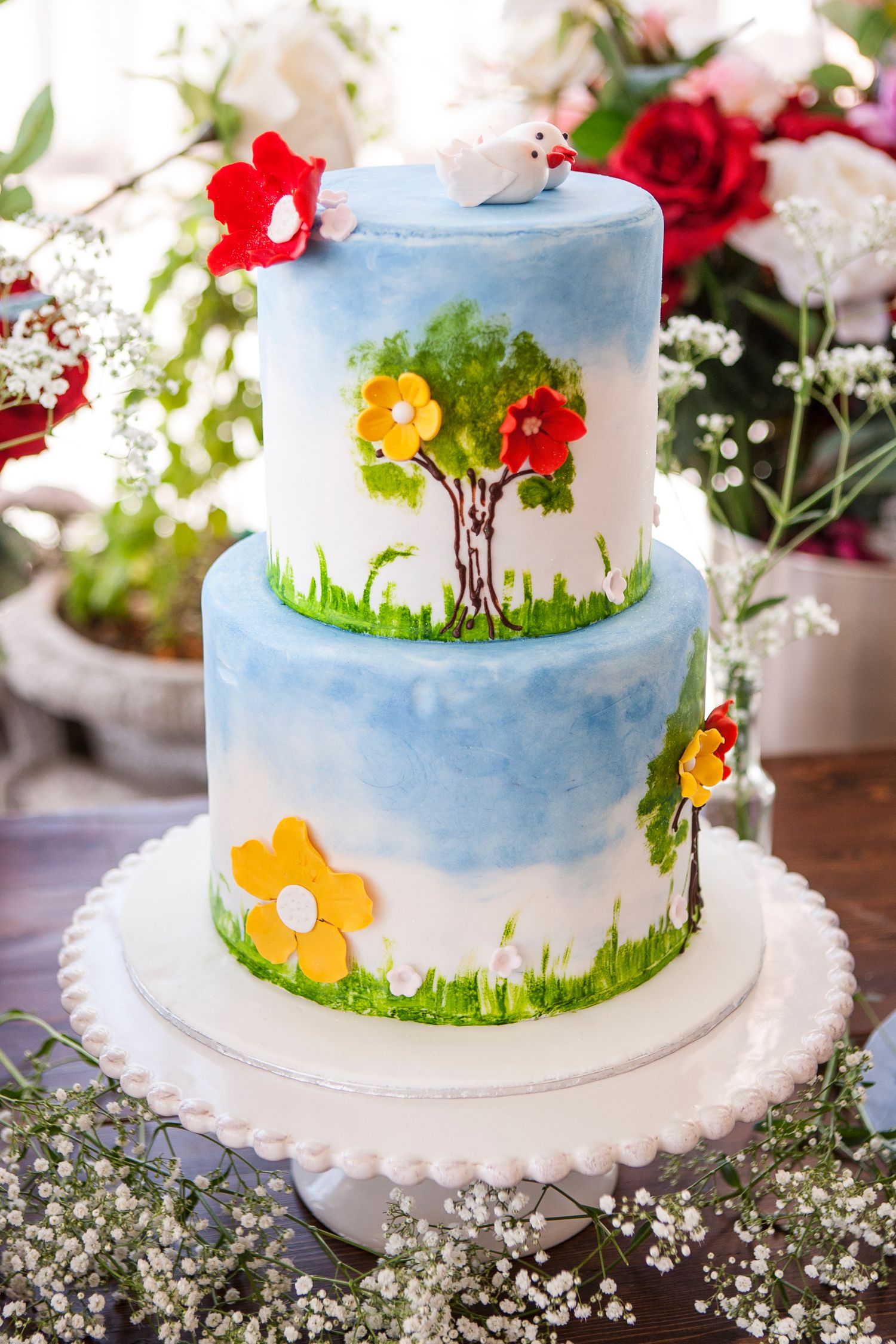 Two-tier blue watercolor wedding cake with painted tree, flowers, and bird topper on white cake stand.
