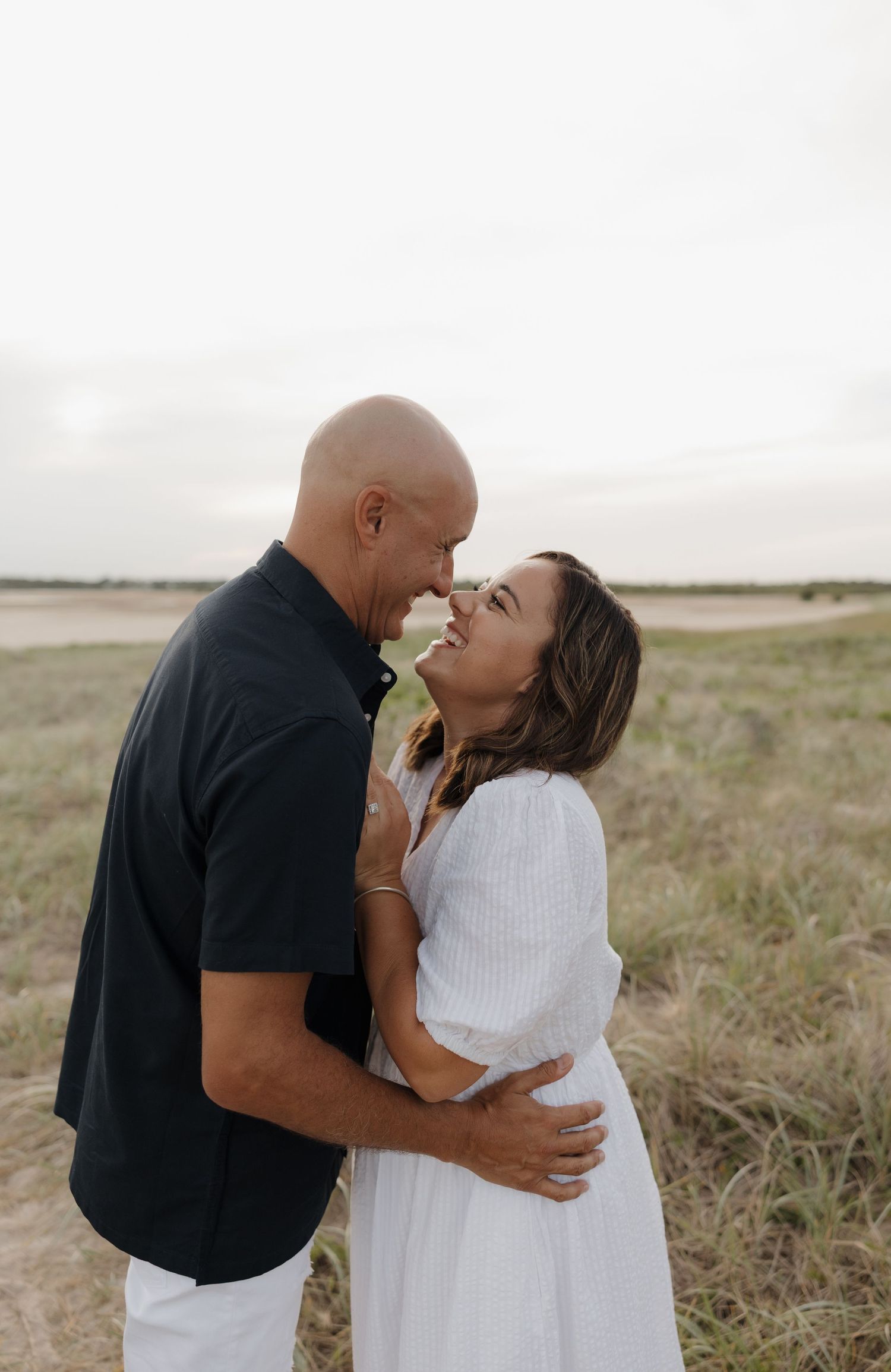 A couple embraces and smiles at each other in an open field, the woman in white dress and man in black shirt. Bundaberg photographer
