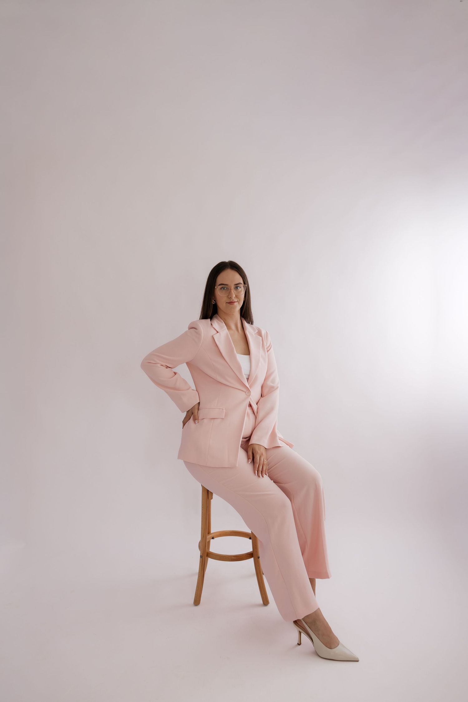 Woman in pink blazer and trousers sitting on wooden stool against white background, wearing cream heels. Bundaberg photographer